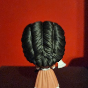 May include: A small figurine viewed from the back, featuring a character with voluminous, dark black hair styled in a central part. The figure wears a long, solid tan-colored dress with a white hemline and holds a small white axe in one hand.