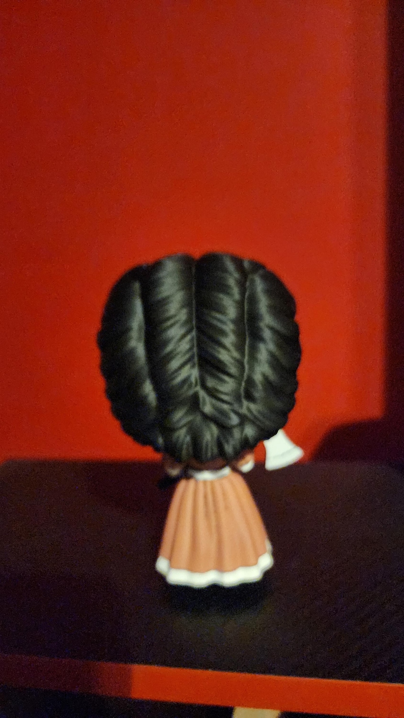 May include: A small figurine viewed from the back, featuring a character with voluminous, dark black hair styled in a central part. The figure wears a long, solid tan-colored dress with a white hemline and holds a small white axe in one hand.