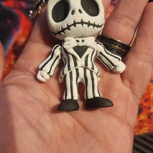 May include: A white and black keychain figure featuring a character with a round skull head, large hollow eyes, and a stitched mouth. The figure wears a pinstriped suit with a bat-shaped bowtie and black shoes, attached to a silver metal keyring.