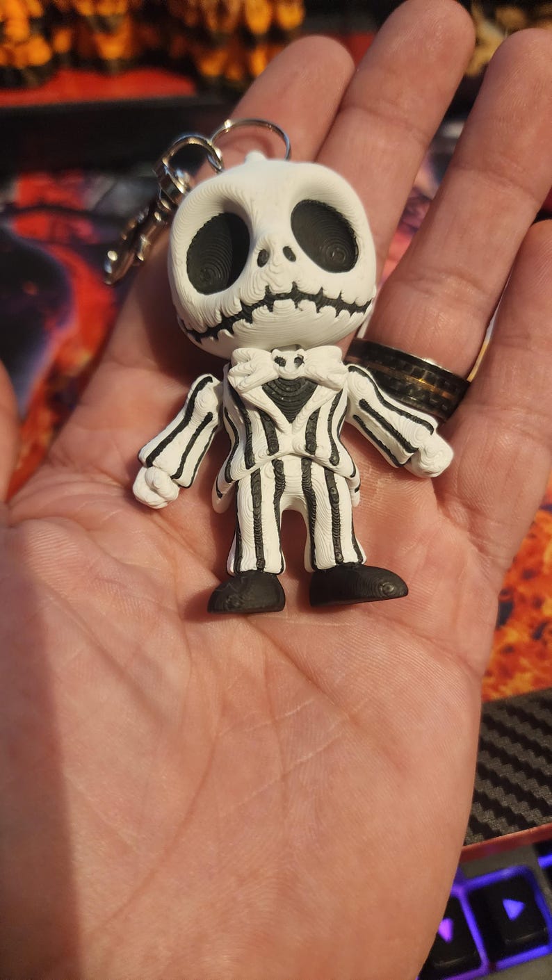 May include: A white and black keychain figure featuring a character with a round skull head, large hollow eyes, and a stitched mouth. The figure wears a pinstriped suit with a bat-shaped bowtie and black shoes, attached to a silver metal keyring.