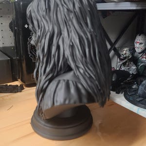 May include: A matte dark gray bust sculpture featuring a figure with long, textured, flowing hair that cascades down the back and shoulders. The figure is mounted on a simple, circular, tiered pedestal base, presented in a monochromatic finish.
