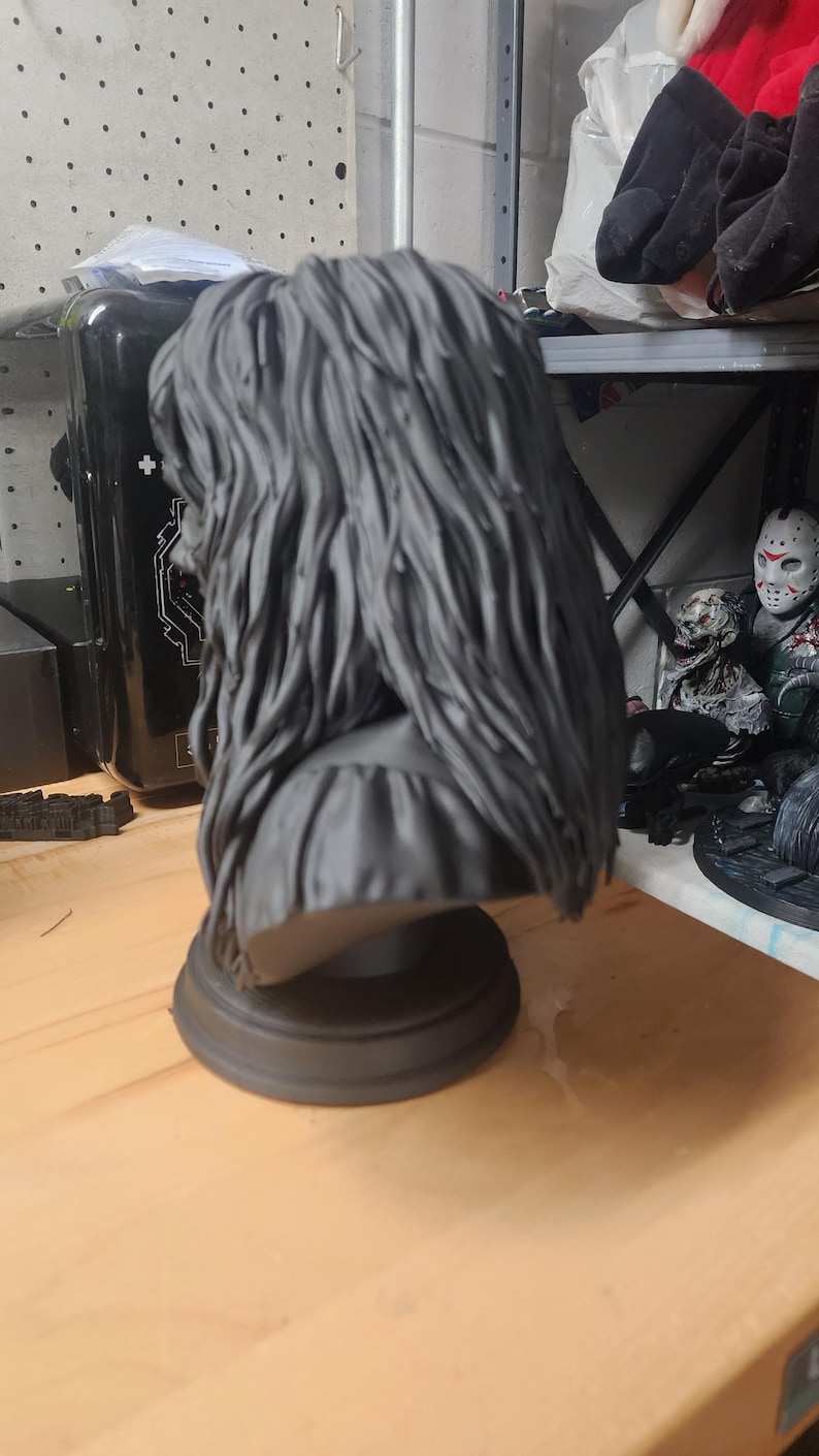May include: A matte dark gray bust sculpture featuring a figure with long, textured, flowing hair that cascades down the back and shoulders. The figure is mounted on a simple, circular, tiered pedestal base, presented in a monochromatic finish.