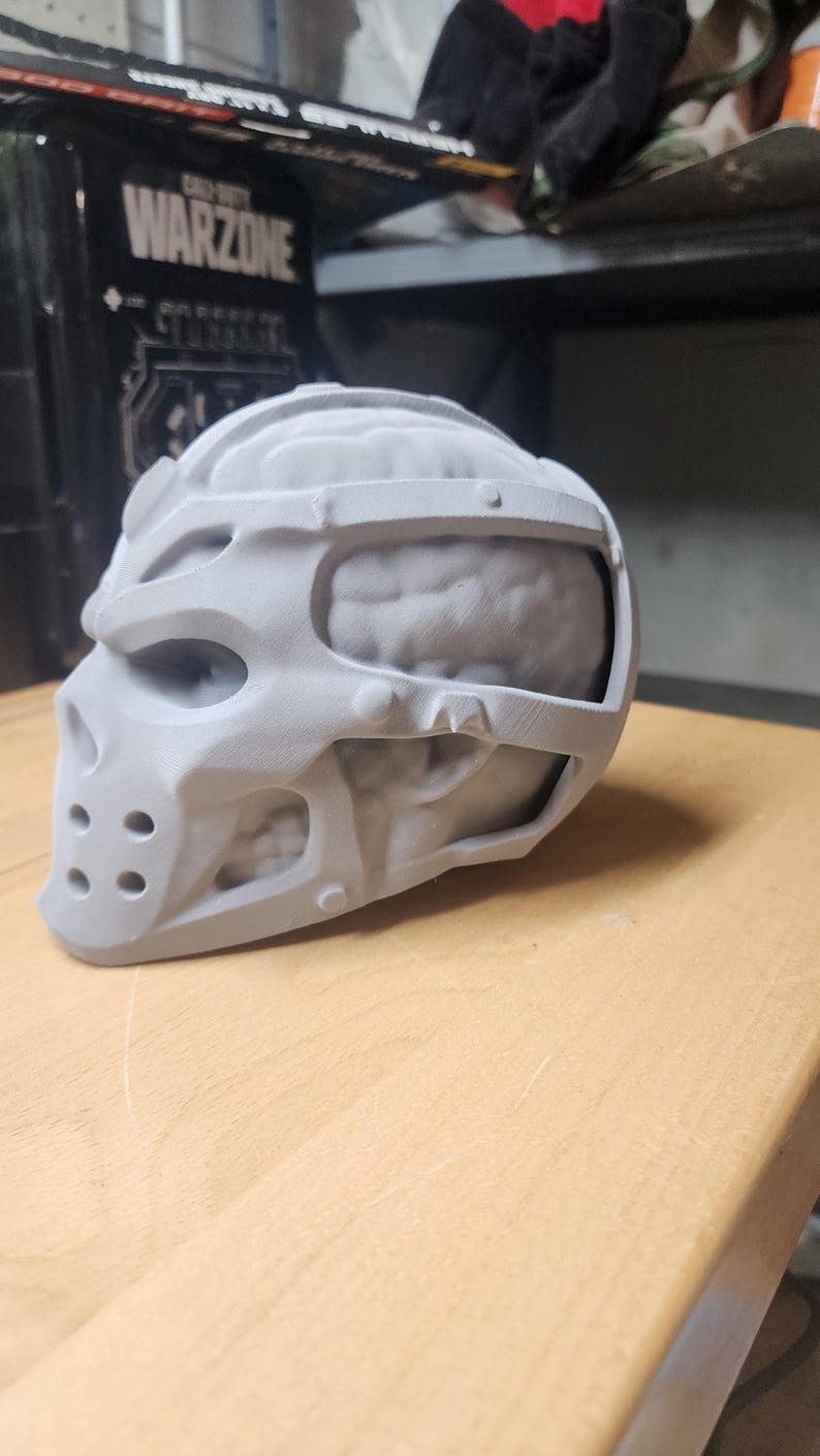 May include: A matte grey 3D printed skull mask featuring a stylized cage-like exterior frame that exposes a textured brain-like structure underneath. The mask includes circular ventilation holes at the chin and a smooth, uniform finish throughout.