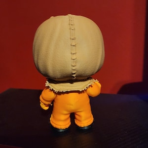 May include: A small vinyl figurine featuring a large, round, tan-colored head resembling a burlap sack with a prominent vertical stitched seam running down the center. The figure wears a bright orange jumpsuit with long sleeves and black shoes.