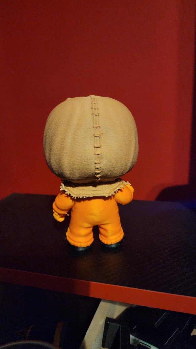 May include: A small vinyl figurine featuring a large, round, tan-colored head resembling a burlap sack with a prominent vertical stitched seam running down the center. The figure wears a bright orange jumpsuit with long sleeves and black shoes.