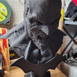 May include: A matte black bust of a character wearing a pointed cowl with a decaying, zombie-like face and exposed teeth. The figure features a bat-shaped emblem on the chest and sits on a base decorated with small skulls and a tombstone design.