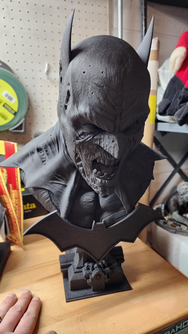 May include: A matte black bust of a character wearing a pointed cowl with a decaying, zombie-like face and exposed teeth. The figure features a bat-shaped emblem on the chest and sits on a base decorated with small skulls and a tombstone design.