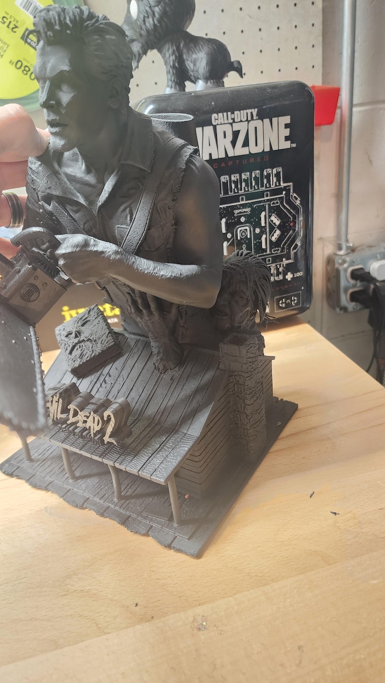 May include: A matte black resin sculpture depicting a character holding a chainsaw, standing on a cabin roof base with the text Evil Dead 2. The figure features a detailed shirt, a book with a face on the cover, and a chimney on the cabin structure.