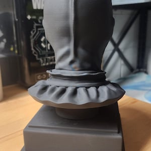 K&ouml;nnte beinhalten: Eine mattschwarze, 3D-gedruckte B&uuml;ste einer Figur mit Zylinder und R&uuml;schenkragen. Die Skulptur ist auf einem quadratischen Sockel montiert und zeigt die R&uuml;ckansicht des Kopfes mit einer vertikalen Nahtlinie, die vom Scheitel bis zum Hals verl&auml;uft.