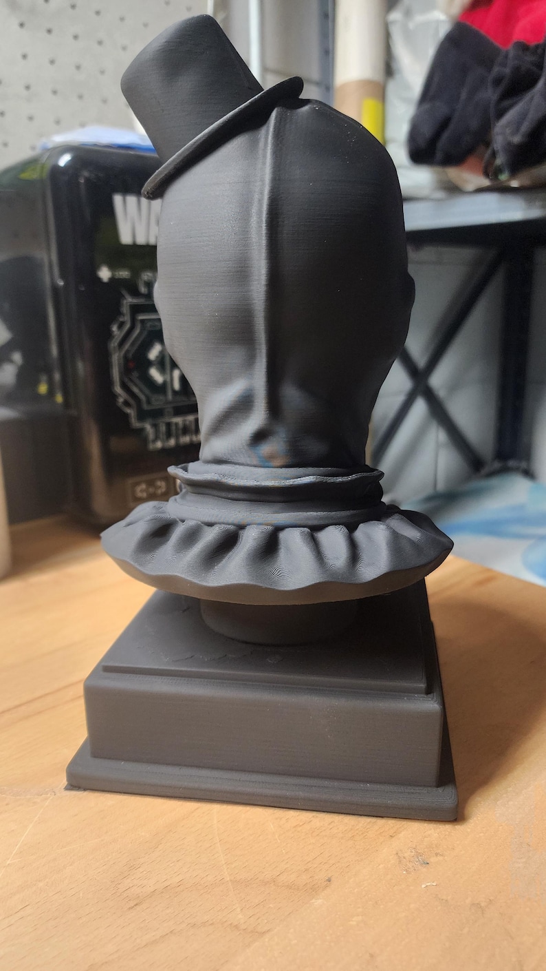 K&ouml;nnte beinhalten: Eine mattschwarze, 3D-gedruckte B&uuml;ste einer Figur mit Zylinder und R&uuml;schenkragen. Die Skulptur ist auf einem quadratischen Sockel montiert und zeigt die R&uuml;ckansicht des Kopfes mit einer vertikalen Nahtlinie, die vom Scheitel bis zum Hals verl&auml;uft.