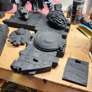 May include: A collection of unpainted black 3D printed miniature terrain pieces, including a brick wall section with a circular hatch, a detached head, a textured circular base, a rectangular tile, and a large creature figure with outstretched arms.
