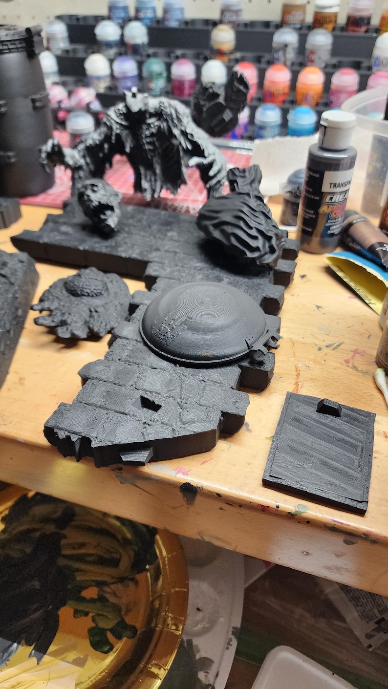 May include: A collection of unpainted black 3D printed miniature terrain pieces, including a brick wall section with a circular hatch, a detached head, a textured circular base, a rectangular tile, and a large creature figure with outstretched arms.
