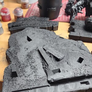 May include: A black 3D printed terrain base featuring a textured rocky surface with rectangular cutouts and raised geometric debris. The piece has a matte finish and displays visible layer lines from the printing process across its surface.