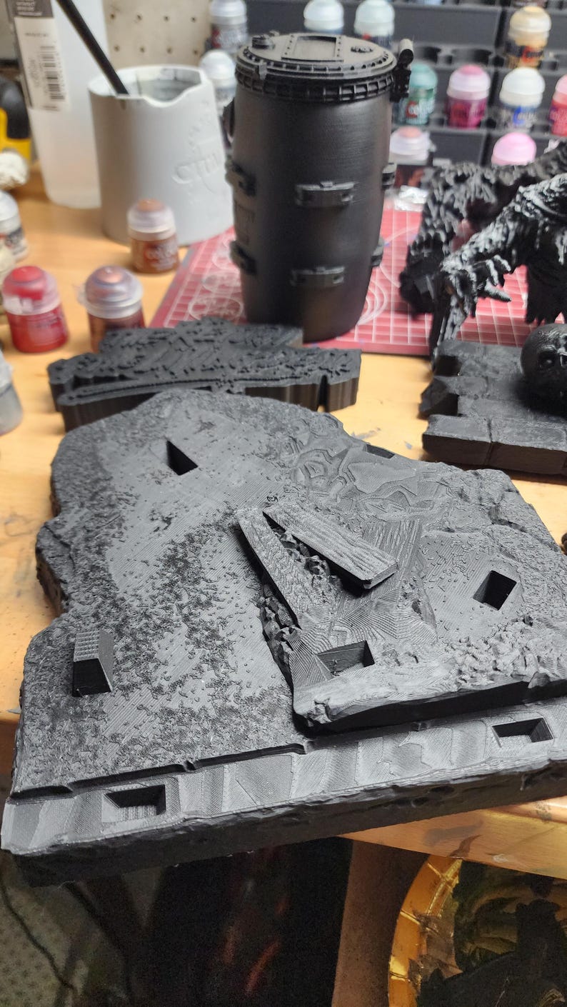 May include: A black 3D printed terrain base featuring a textured rocky surface with rectangular cutouts and raised geometric debris. The piece has a matte finish and displays visible layer lines from the printing process across its surface.
