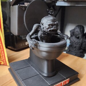 May include: A matte black 3D printed figurine featuring a creature emerging from a toilet bowl, with a second creature perched on the lid. The base is square and displays the word GHOULIES in red and white lettering on the front edge.