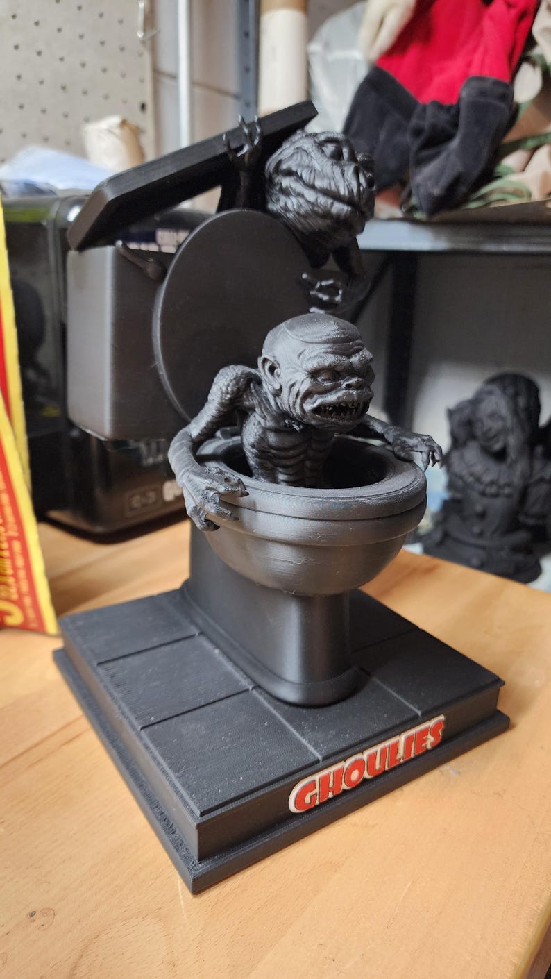 May include: A matte black 3D printed figurine featuring a creature emerging from a toilet bowl, with a second creature perched on the lid. The base is square and displays the word GHOULIES in red and white lettering on the front edge.