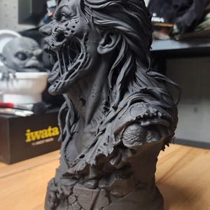 May include: A matte black 3D printed bust of a creature with long flowing hair, a decaying face, and an exposed jaw structure. The figure features a tattered garment on the shoulders and rests on a circular base with a cracked stone texture.