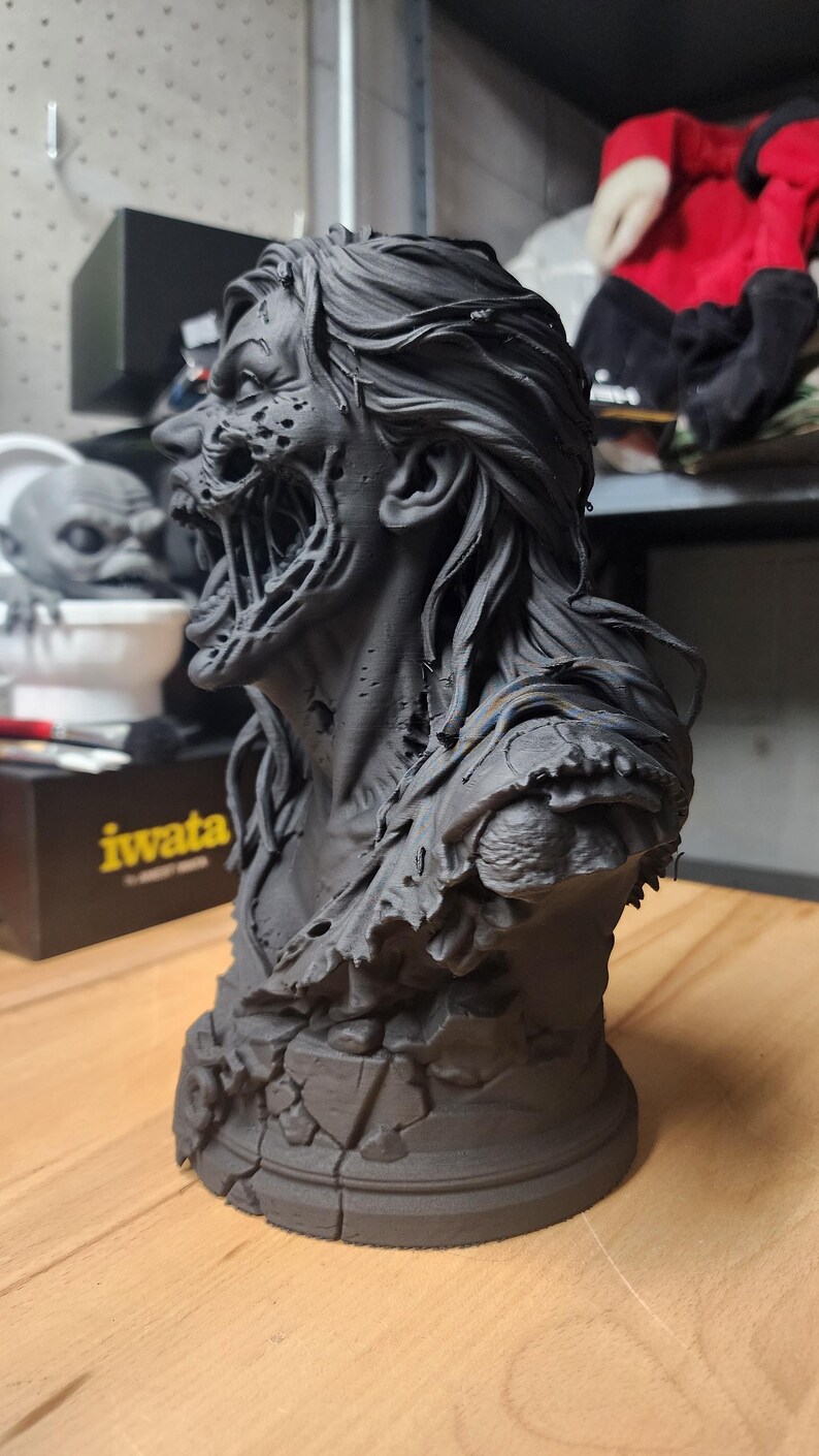 May include: A matte black 3D printed bust of a creature with long flowing hair, a decaying face, and an exposed jaw structure. The figure features a tattered garment on the shoulders and rests on a circular base with a cracked stone texture.