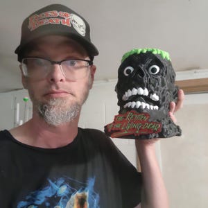 May include: A black, sculpted head figurine featuring a textured surface, white eyes and teeth, and bright green hair. The base of the figurine displays the red and green text "THE RETURN OF THE LIVING DEAD" in a stylized, jagged font.