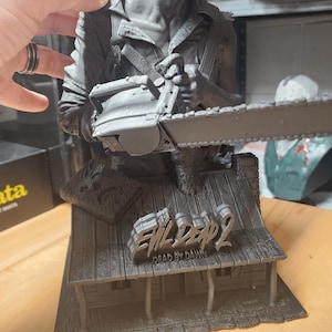 May include: A gray monochromatic resin bust of a character holding a chainsaw, mounted on a base shaped like a cabin roof. The base features the text Evil Dead 2 Dead by Dawn and a small book. The figure has a detailed facial expression and clothing.