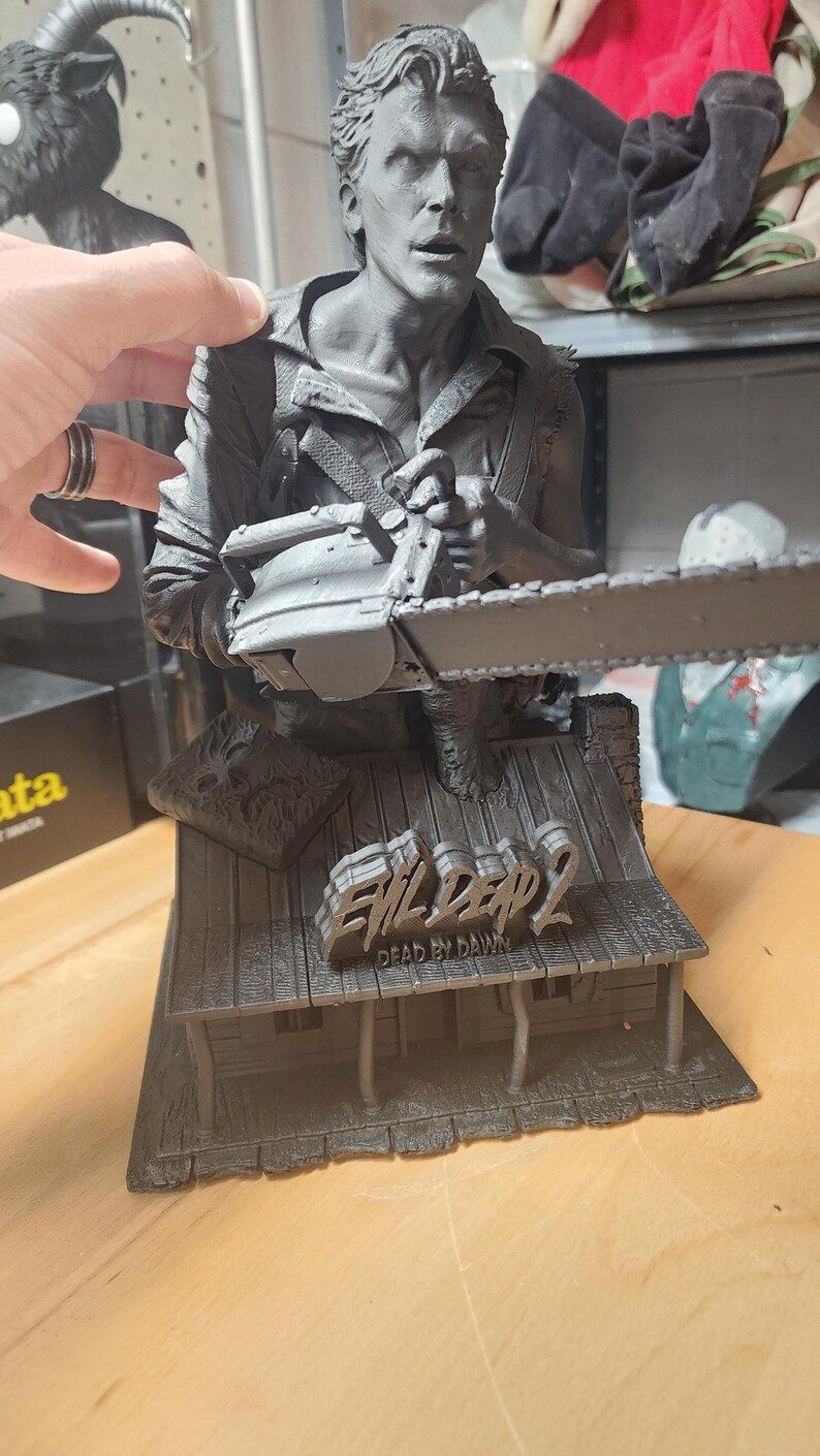 May include: A gray monochromatic resin bust of a character holding a chainsaw, mounted on a base shaped like a cabin roof. The base features the text Evil Dead 2 Dead by Dawn and a small book. The figure has a detailed facial expression and clothing.