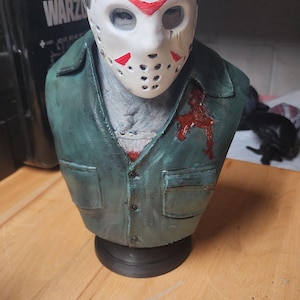 May include: A sculpted bust of a figure wearing a white hockey mask with red triangular markings and a dark green collared shirt. The shirt features a prominent red blood-like stain on the left chest area. The figure has a grey textured neck.