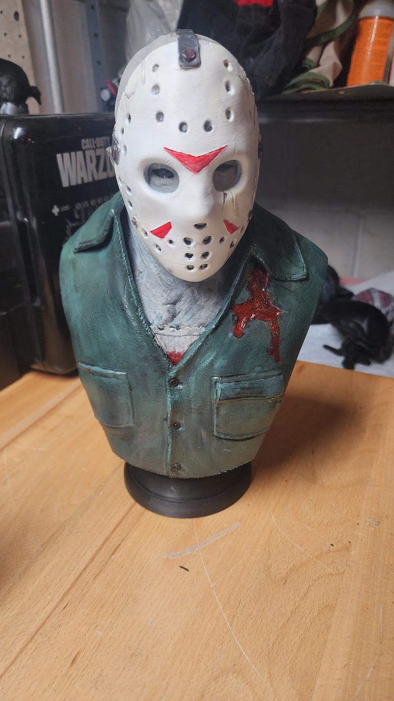 May include: A sculpted bust of a figure wearing a white hockey mask with red triangular markings and a dark green collared shirt. The shirt features a prominent red blood-like stain on the left chest area. The figure has a grey textured neck.