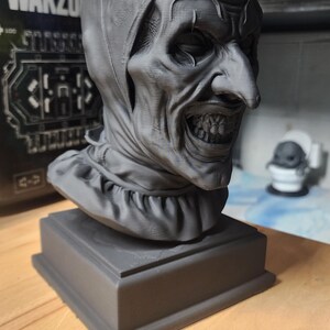 K&ouml;nnte beinhalten: Eine matte graue 3D-gedruckte B&uuml;ste einer Clownfigur mit eng anliegender Kapuze, ger&uuml;schtem Kragen, markanter spitzer Nase und einem breiten, zahnigen Grinsen. Die Skulptur ist auf einem quadratischen Sockel montiert und zeigt sichtbare Druckschichten.