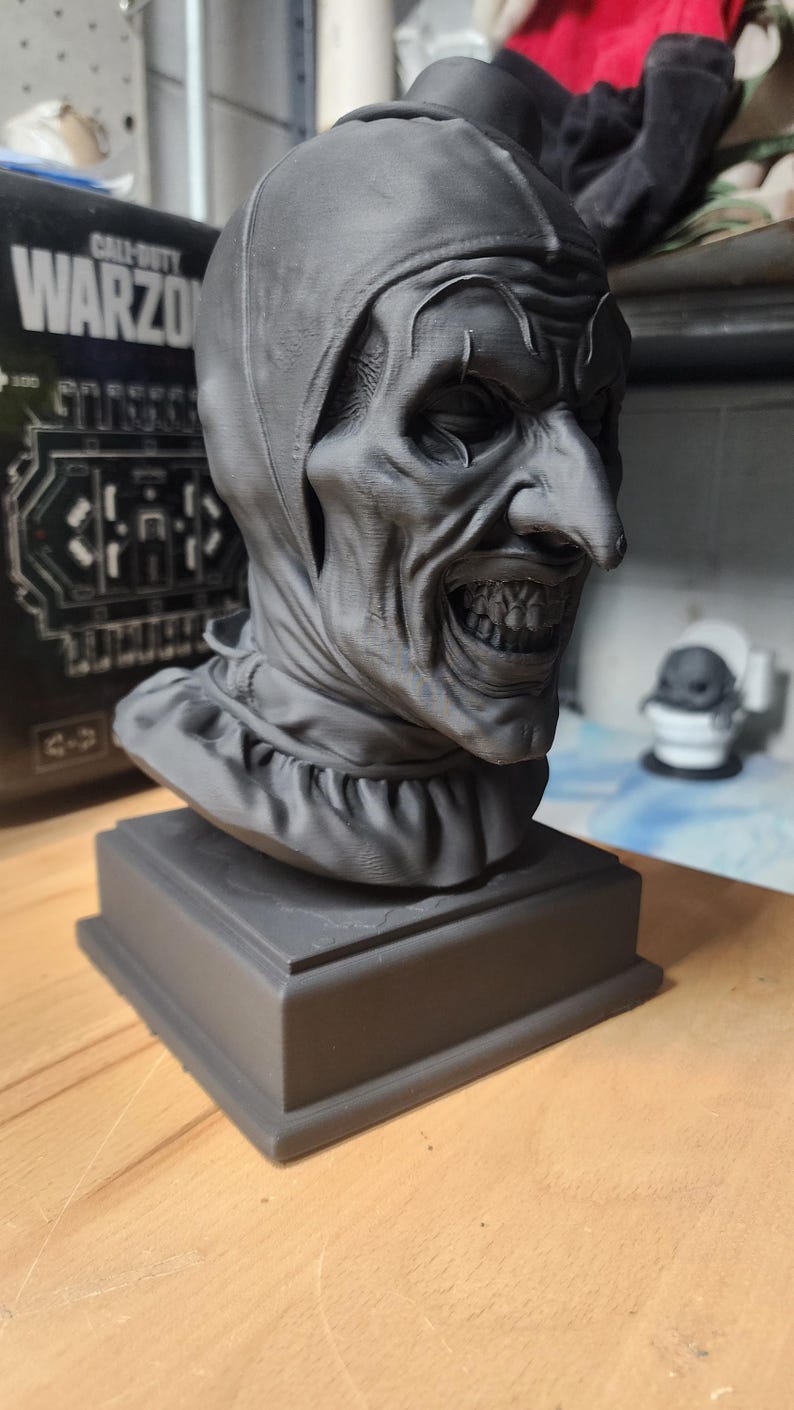 K&ouml;nnte beinhalten: Eine matte graue 3D-gedruckte B&uuml;ste einer Clownfigur mit eng anliegender Kapuze, ger&uuml;schtem Kragen, markanter spitzer Nase und einem breiten, zahnigen Grinsen. Die Skulptur ist auf einem quadratischen Sockel montiert und zeigt sichtbare Druckschichten.