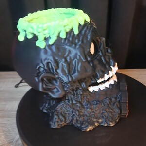May include: A black skull-shaped container with a textured, melted-look surface. The top rim features a bright green, dripping slime effect. The skull has white teeth and a hollow interior, presented as a decorative vessel or holder for small items.