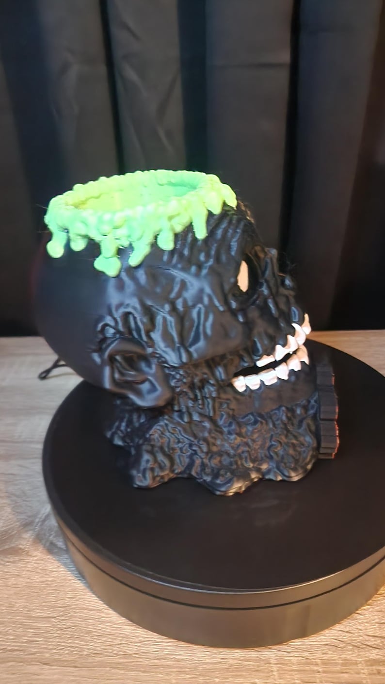 May include: A black skull-shaped container with a textured, melted-look surface. The top rim features a bright green, dripping slime effect. The skull has white teeth and a hollow interior, presented as a decorative vessel or holder for small items.