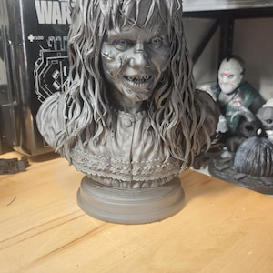 May include: A monochromatic gray bust of a person with long, wavy, unkempt hair and a distressed facial expression. The figure wears a collared shirt with a buttoned front and a detailed lace-like trim along the hem, all set on a circular base.