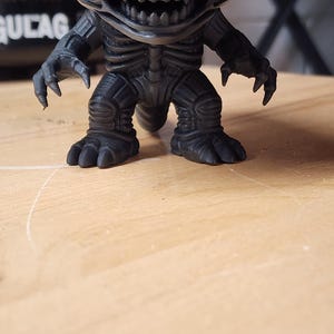 May include: A matte black vinyl figure of the Xenomorph creature from the Alien franchise. The figure features a large elongated head, a wide mouth with visible sharp teeth, clawed hands, and a textured, segmented body with a tail.