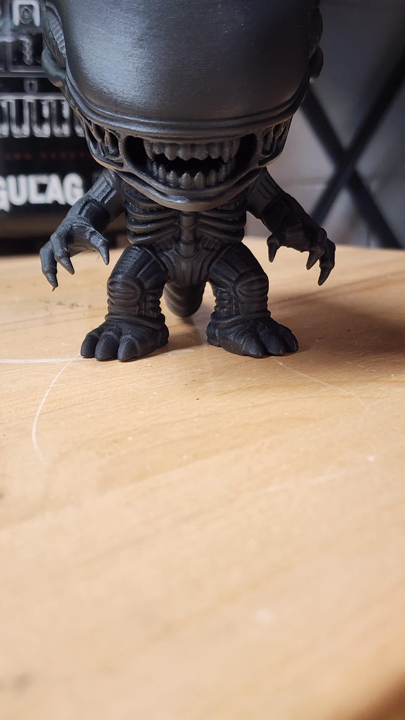 May include: A matte black vinyl figure of the Xenomorph creature from the Alien franchise. The figure features a large elongated head, a wide mouth with visible sharp teeth, clawed hands, and a textured, segmented body with a tail.