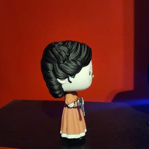 May include: A stylized vinyl collectible figure featuring a character with dark, textured hair styled in an updo, wearing a peach-colored dress with white trim at the sleeves and hem, and holding a small silver-colored object in one hand.