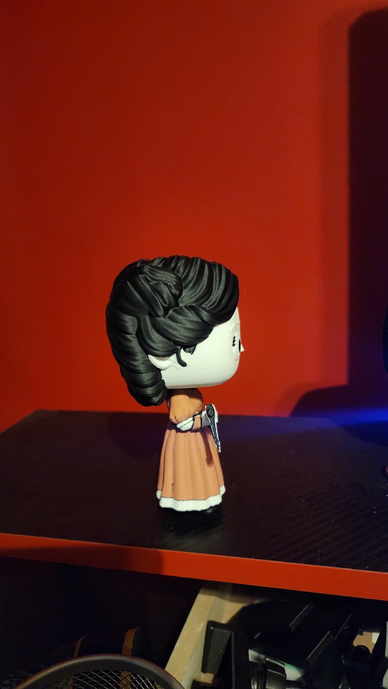 May include: A stylized vinyl collectible figure featuring a character with dark, textured hair styled in an updo, wearing a peach-colored dress with white trim at the sleeves and hem, and holding a small silver-colored object in one hand.