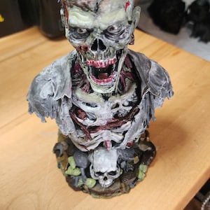 Op de afbeelding: Een gedetailleerde zombiebuste met een bleek, vergaan gezicht met blootliggende tanden, een wijd open mond en zichtbaar spierweefsel. Het figuur draagt een gescheurd grijs hemd over een blootliggende borstkas op een voetstuk met een schedel.