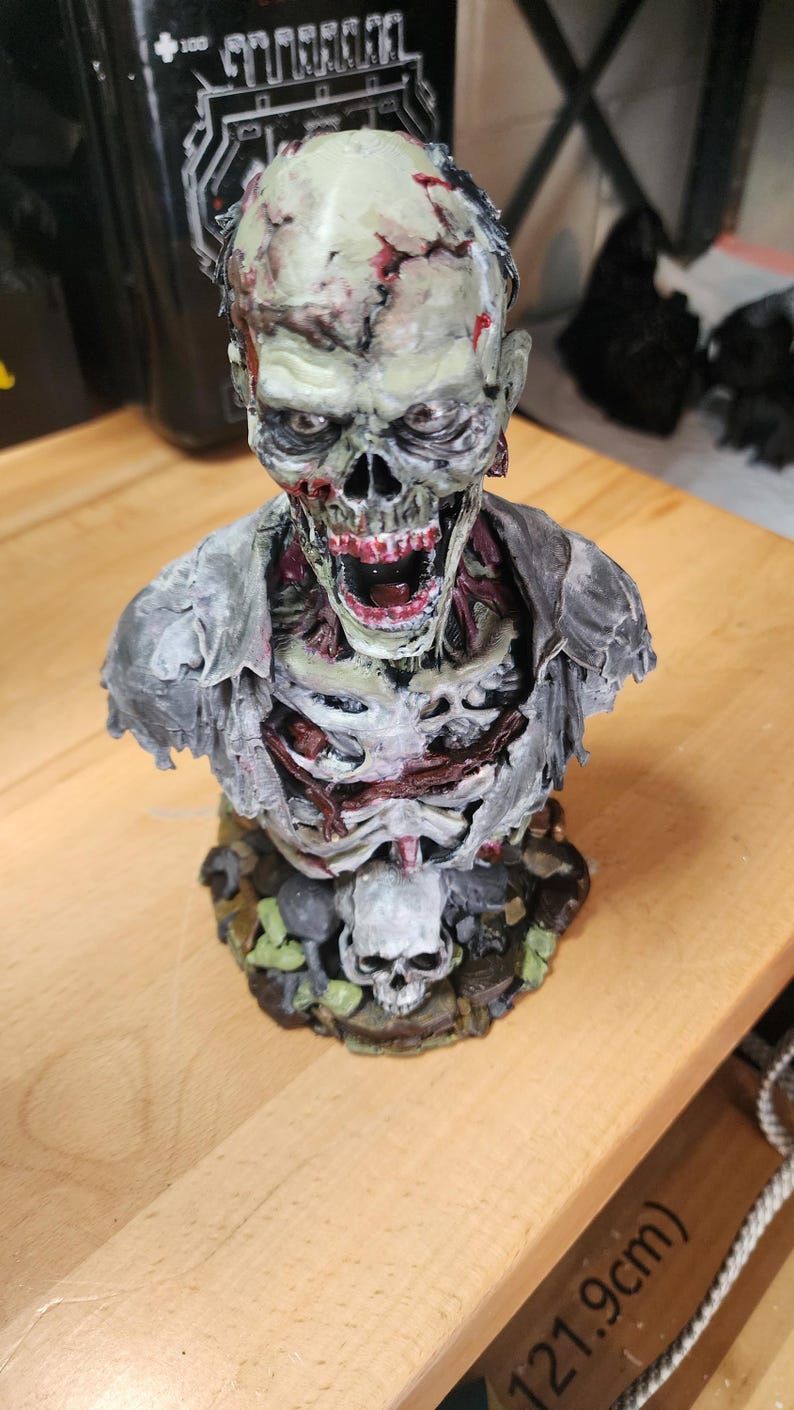 Op de afbeelding: Een gedetailleerde zombiebuste met een bleek, vergaan gezicht met blootliggende tanden, een wijd open mond en zichtbaar spierweefsel. Het figuur draagt een gescheurd grijs hemd over een blootliggende borstkas op een voetstuk met een schedel.
