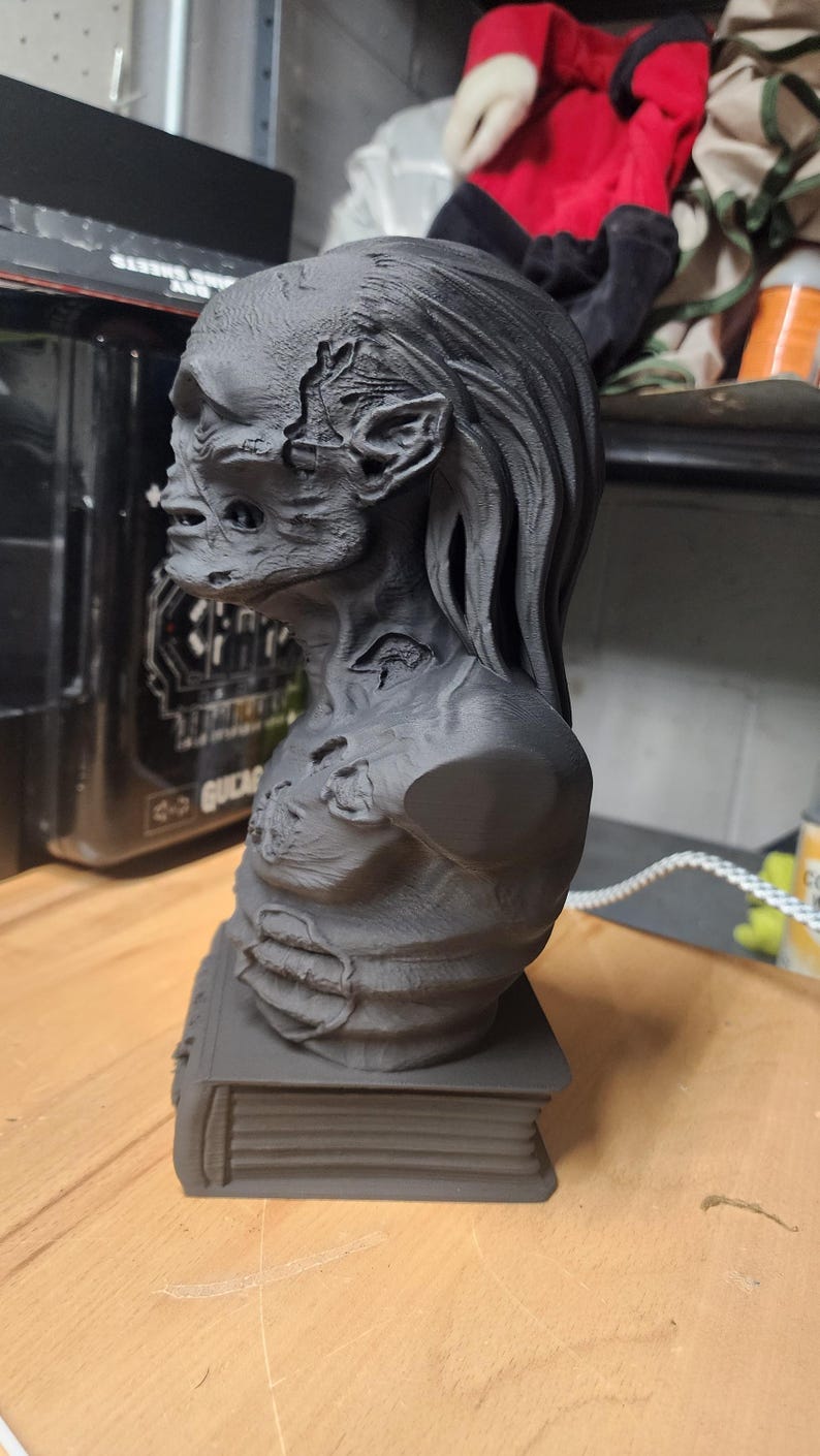 May include: A matte black 3D printed bust of a creature with textured, weathered skin, pointed ears, and long hair, mounted on a rectangular base shaped like a closed book. The figure displays detailed facial features and visible surface imperfections.
