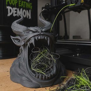 May include: A matte dark gray 3D printed demon bust with large horns and an open mouth filled with tangled black and neon green plastic filament waste. A sign behind the sculpture reads FILAMENT POOP EATING DEMON in white and green text.