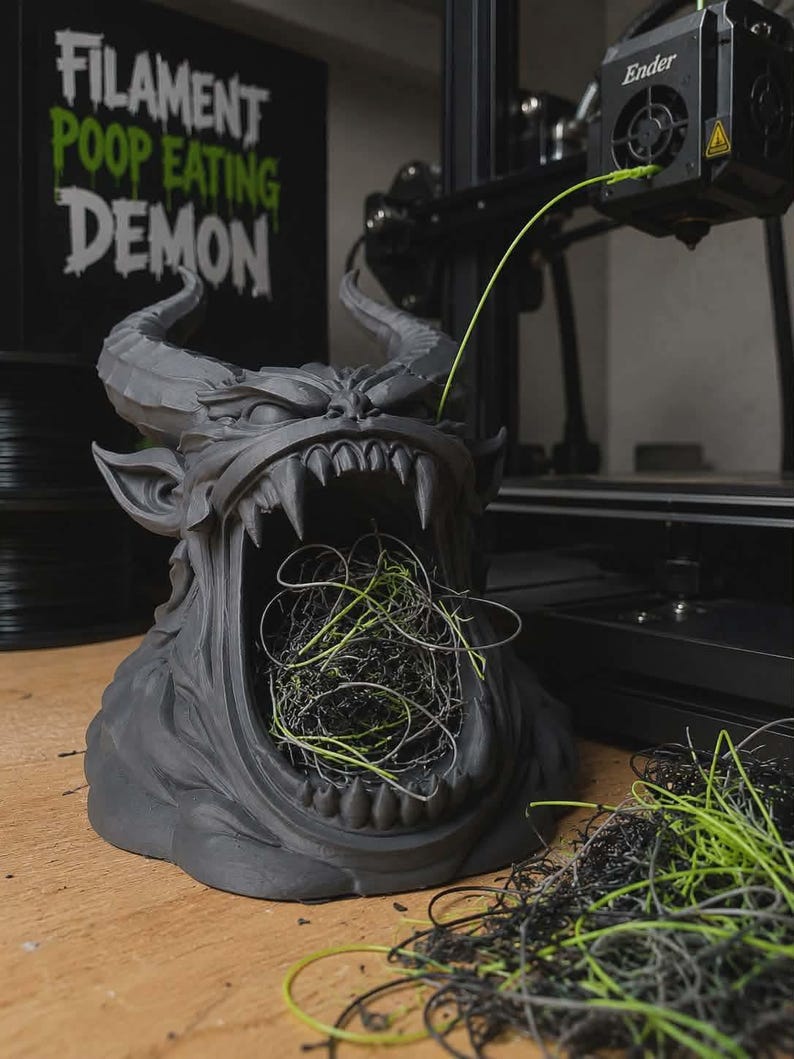 May include: A matte dark gray 3D printed demon bust with large horns and an open mouth filled with tangled black and neon green plastic filament waste. A sign behind the sculpture reads FILAMENT POOP EATING DEMON in white and green text.