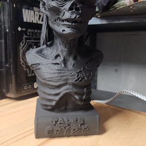 May include: A matte black 3D printed bust of the Crypt Keeper character, featuring a skeletal face with deep-set eyes, a prominent brow, and a textured chest. The base of the figure displays the embossed text TALES FROM THE CRYPT in capital letters.