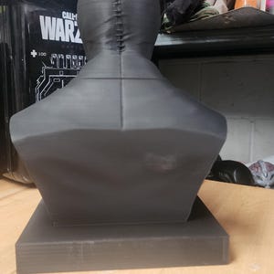 May include: A matte black 3D printed bust of a figure wearing a tight-fitting mask with a visible vertical seam running from the crown of the head down the back of the neck, mounted on a square base, viewed from the rear perspective.