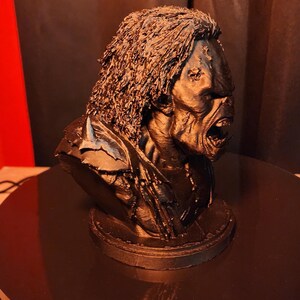 May include: A black 3D printed bust of a creature with long, textured hair, a prominent brow, and an open mouth revealing teeth. The figure features detailed facial skin texture and is mounted on a circular base, captured from a side profile view.