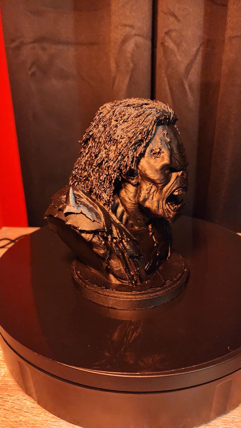 May include: A black 3D printed bust of a creature with long, textured hair, a prominent brow, and an open mouth revealing teeth. The figure features detailed facial skin texture and is mounted on a circular base, captured from a side profile view.
