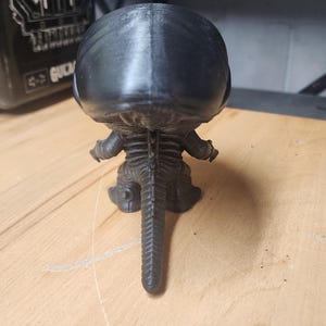May include: A black 3D printed figurine of a creature with a large smooth head, a segmented spine, and a long pointed tail. The figure is shown from the back, displaying a textured surface and a distinct vertical seam running down the center of the head.