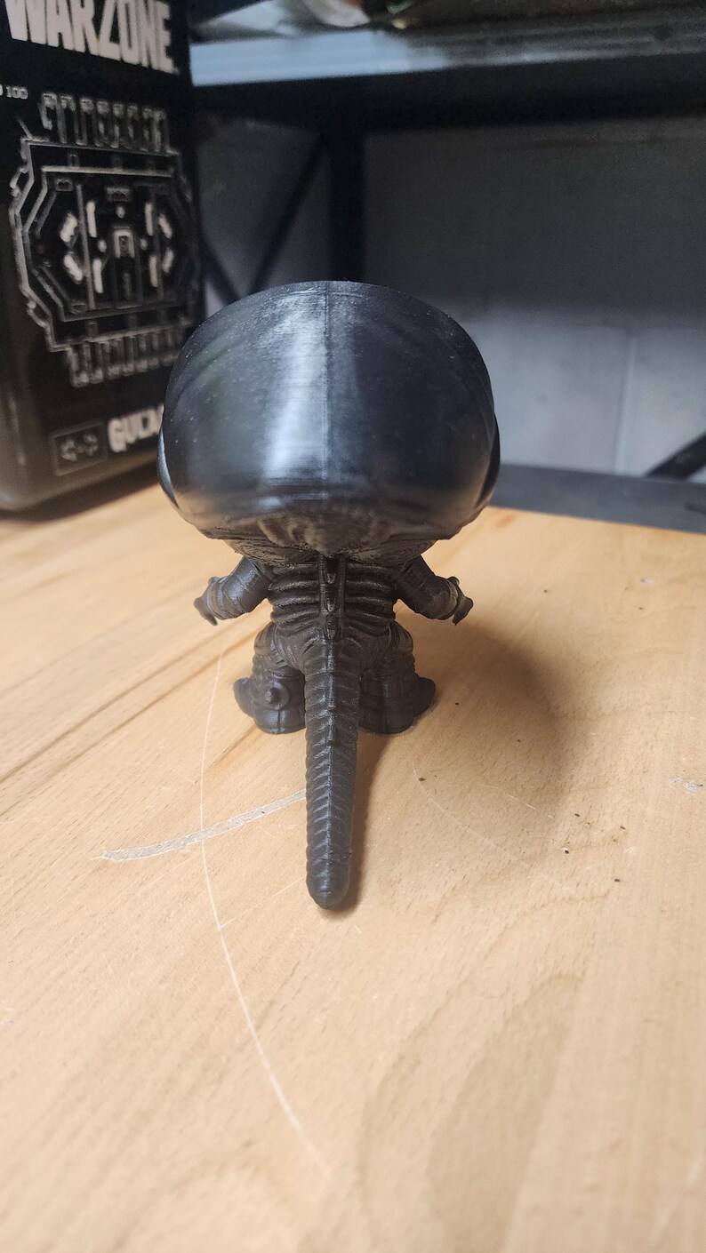 May include: A black 3D printed figurine of a creature with a large smooth head, a segmented spine, and a long pointed tail. The figure is shown from the back, displaying a textured surface and a distinct vertical seam running down the center of the head.