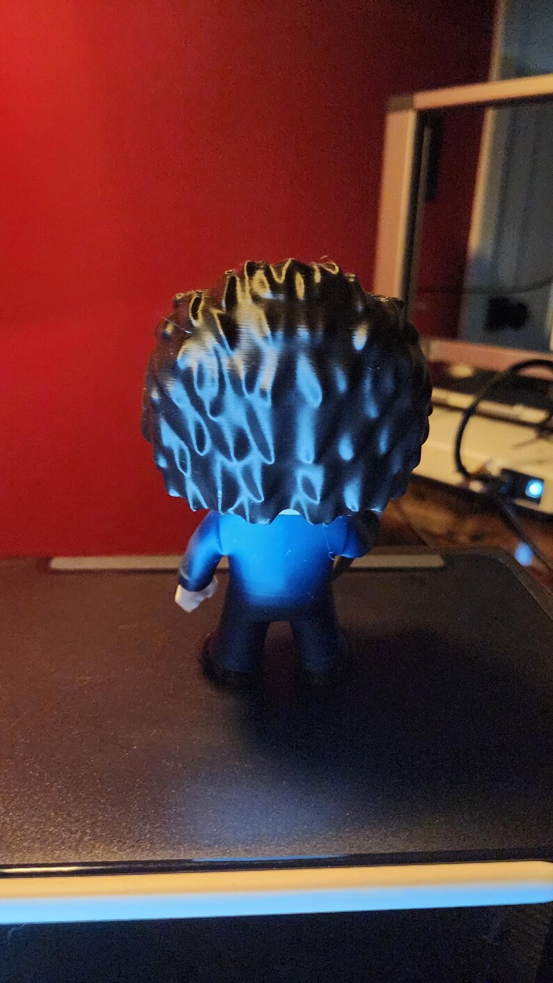 May include: A small stylized figurine viewed from the back, featuring a large head with textured black hair, a blue jacket, and black trousers. The figure has a smooth finish and stands upright, showcasing a simple design with distinct color blocking.
