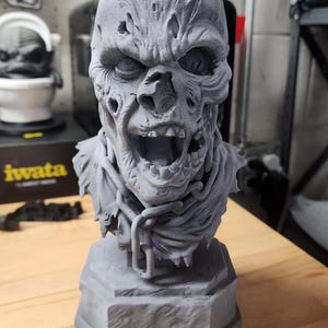 May include: A gray 3D printed bust of a decaying zombie figure with a wide-open mouth, exposed teeth, and textured skin. The figure features a tattered collar and a chain draped around the neck, all mounted on a solid hexagonal base.