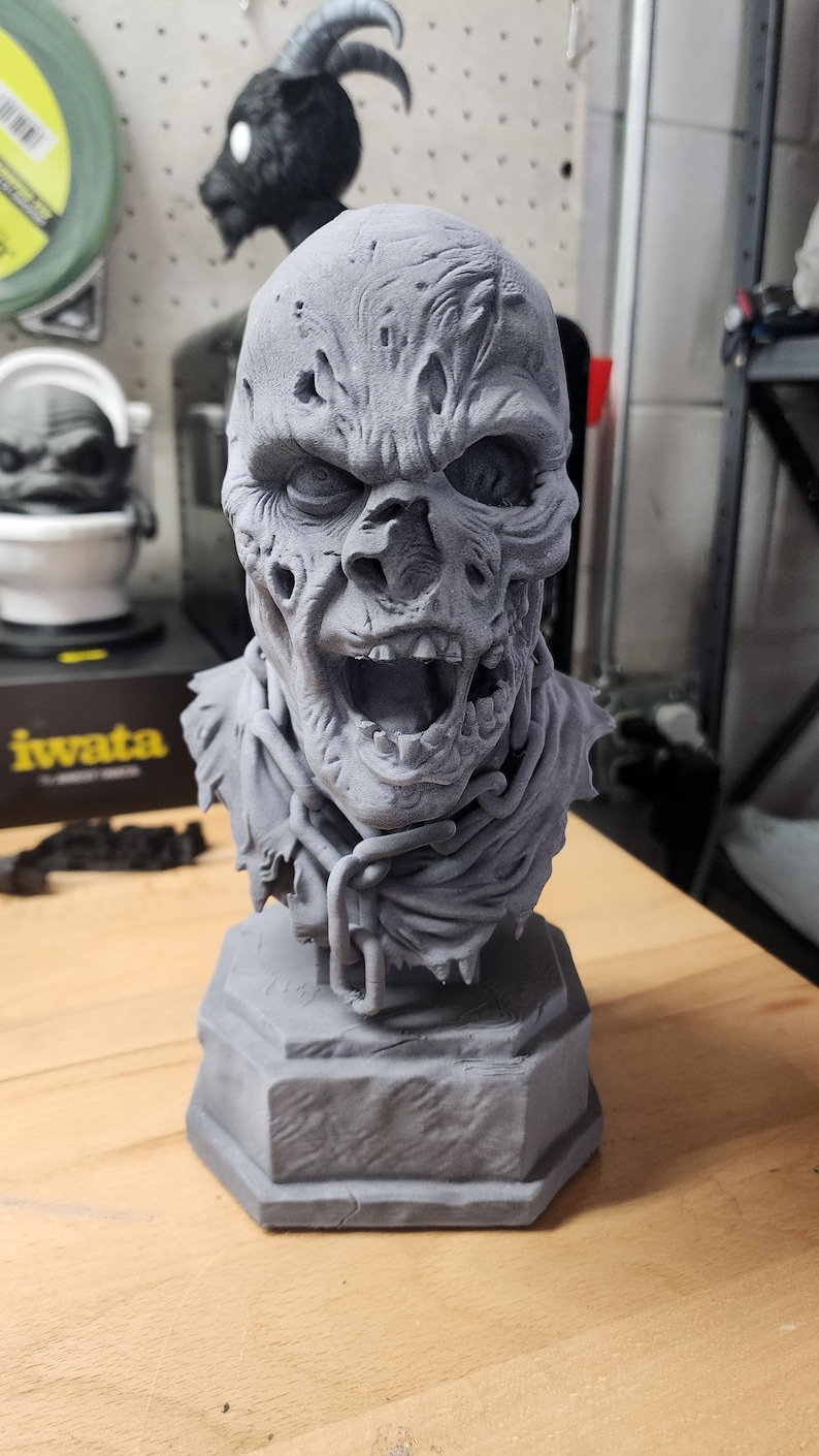 May include: A gray 3D printed bust of a decaying zombie figure with a wide-open mouth, exposed teeth, and textured skin. The figure features a tattered collar and a chain draped around the neck, all mounted on a solid hexagonal base.